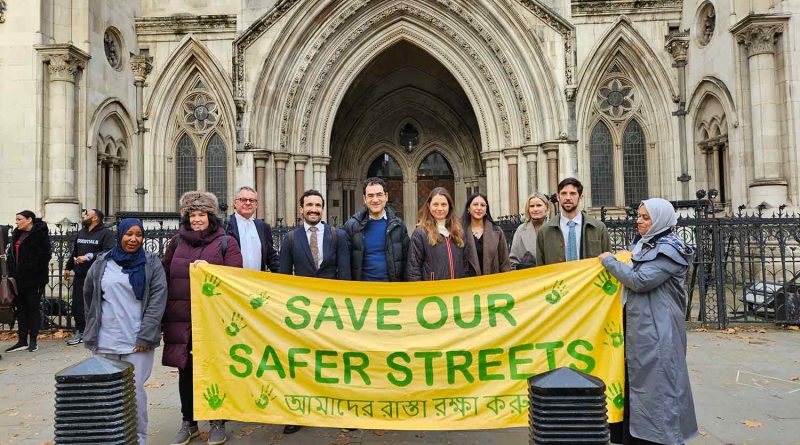 Photo of campaigners in front of the Royal Courts of Justice, holding a yellow banner with green lettering which spells 'Save Our Safer Streets' in English and in Bengali.